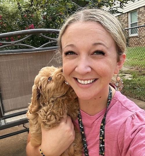 A close-up of a white woman with blonde hair. She's looking into the camera and smiling while holding a small poodle-mix dog that is licking her face.