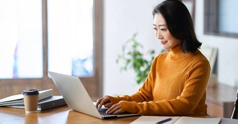 asian woman happily using laptop on table with coffee and files at workspace