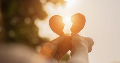 A pair of hands holding up two pieces of a broken heart toward the setting sun. 