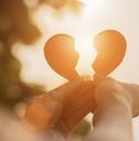A pair of hands holding up two pieces of a broken heart toward the setting sun. 