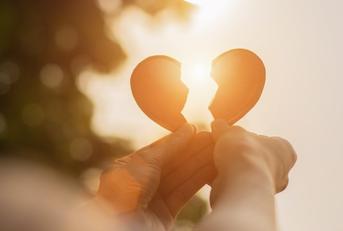A pair of hands holding up two pieces of a broken heart toward the setting sun. 