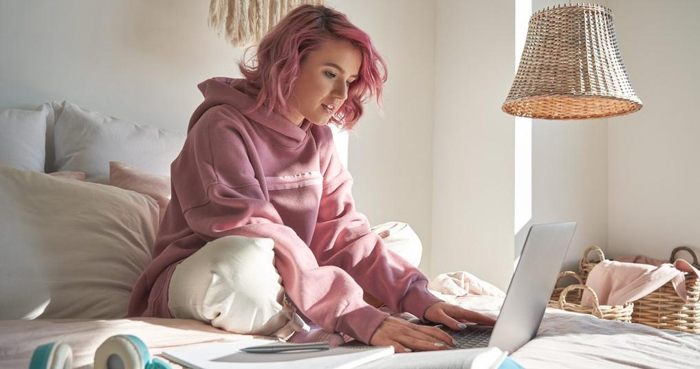 A young woman sits cross-legged on a bed and looks at a laptop screen in front of her. 