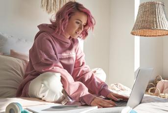 A young woman sits cross-legged on a bed and looks at a laptop screen in front of her. 