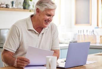 An older man sits in front of a laptop. He holds a piece of paper in one hand and he is smiling at an unseen image on the laptop screen.