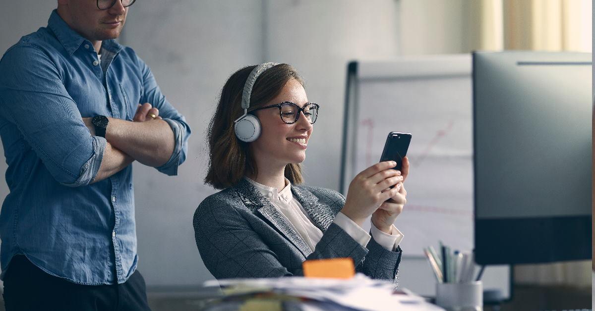 Happy woman using smartphone at office with boss standing behind her