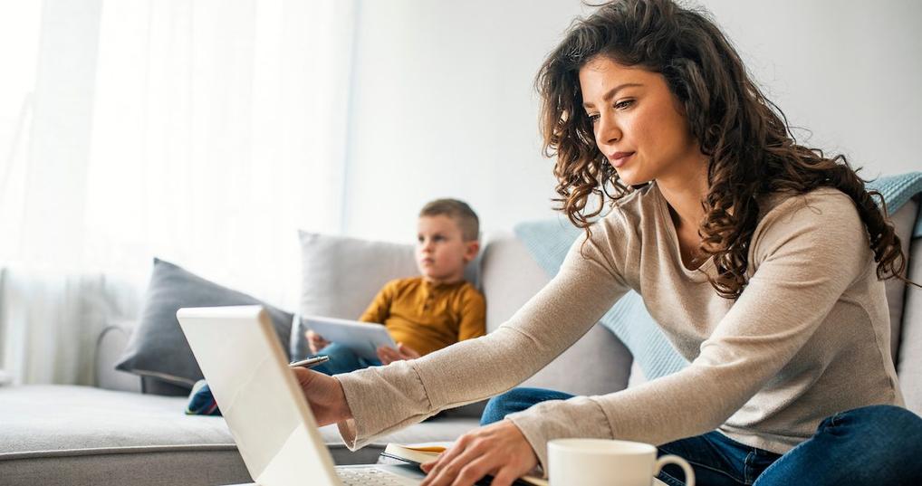 A mom works from her laptop while sitting on the couch next to a young child. 