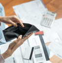 A woman opens an empty black wallet, while bills and paperwork scatter the floor.